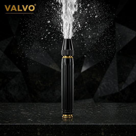 A VALVO black and gold handheld water spray nozzle with a powerful water jet spraying upward, set on a dark stone surface against a dark faceted background.